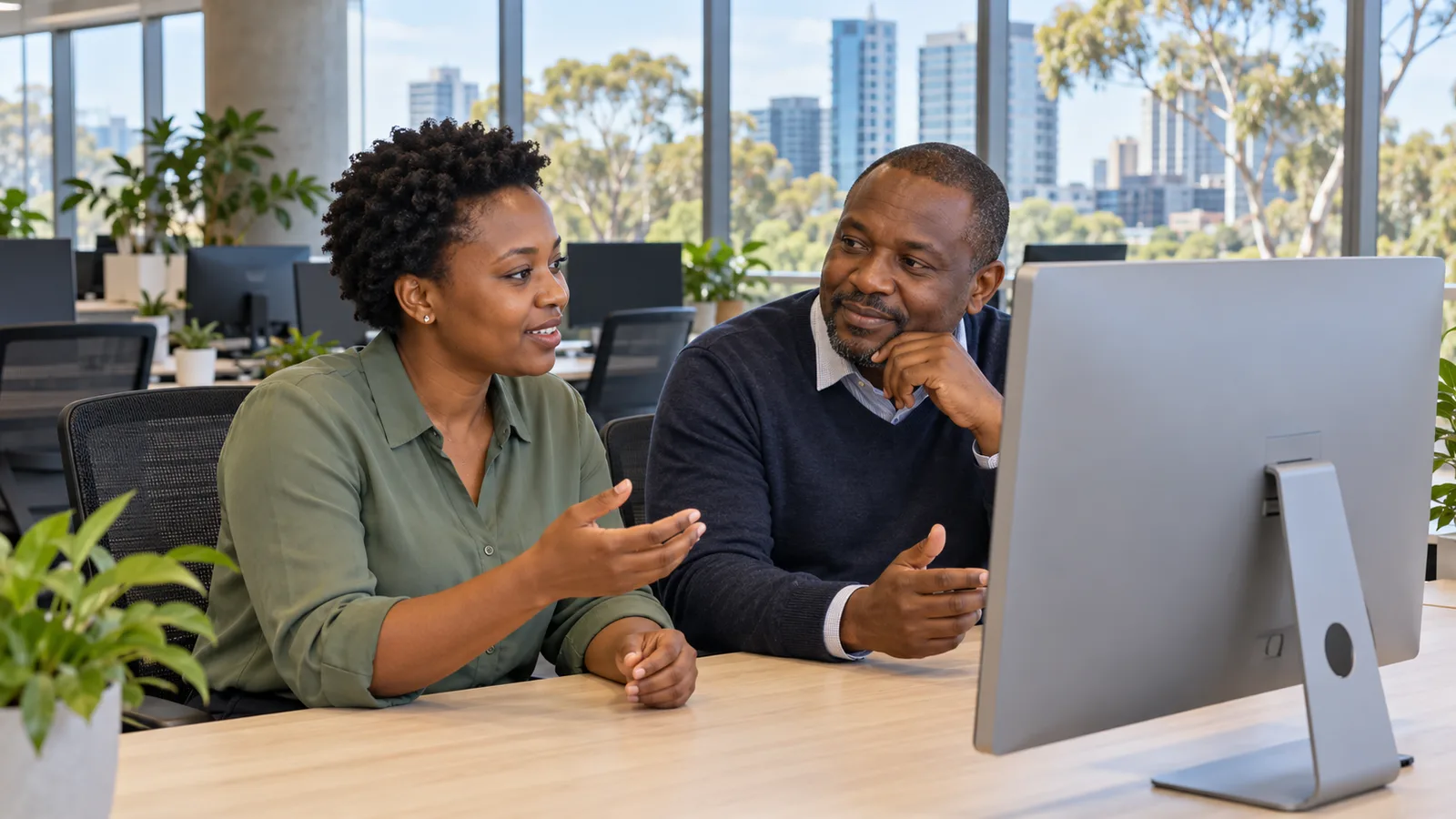 Two Tanzanian colleagues discuss work beside a desktop computer in a bright Australian office.